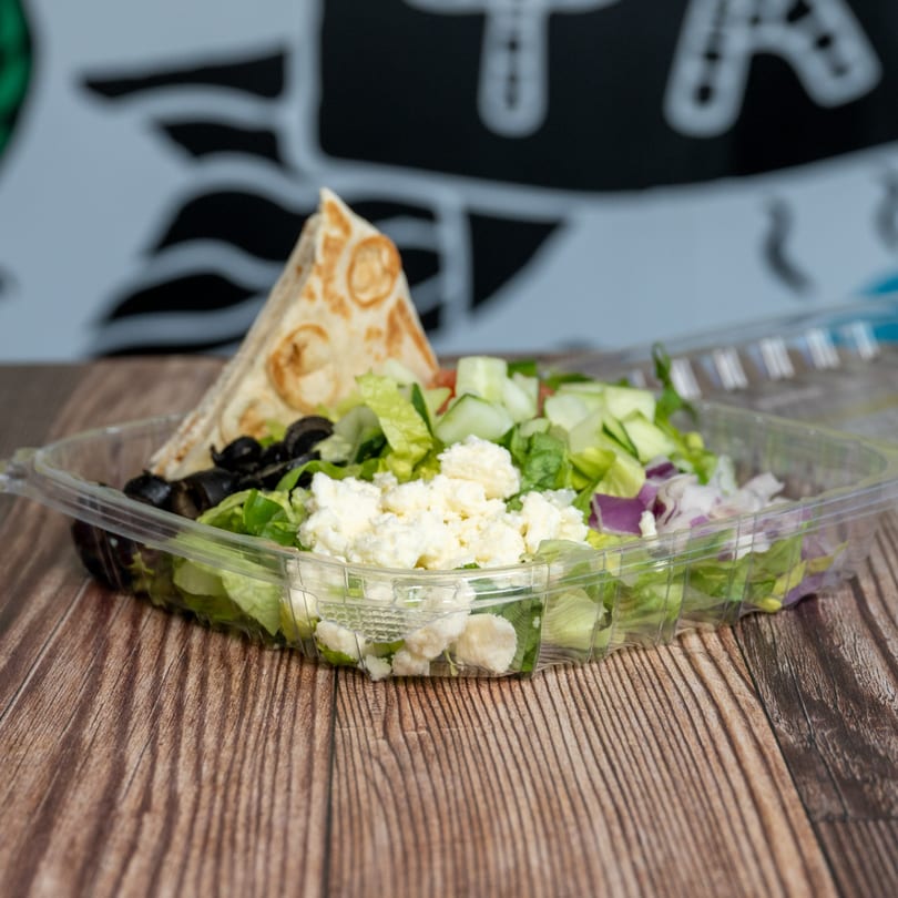 Natural Greens salad with lettuce, feta, olives, cucumbers, red onion, and pita bread in a plastic container.