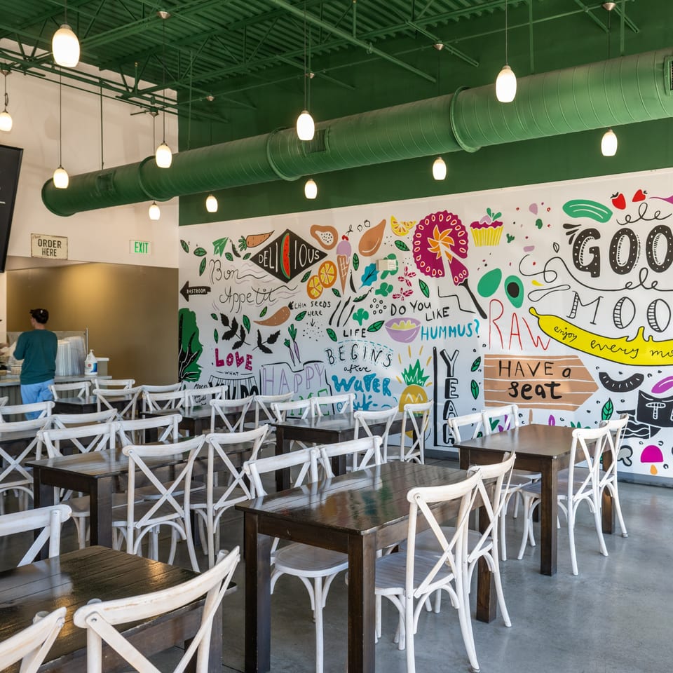 Interior of Natural Greens restaurant with dark wooden tables, white chairs, and a colorful food-themed mural.