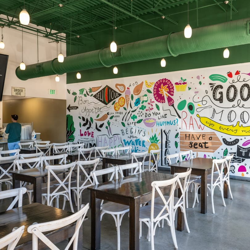 Interior of Natural Greens restaurant with dark wooden tables, white chairs, and a colorful food-themed mural.