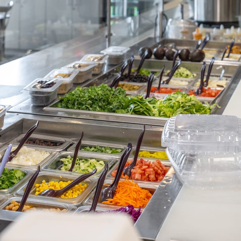 A salad bar at Natural Greens with various fresh ingredients in metal bins and serving tongs.