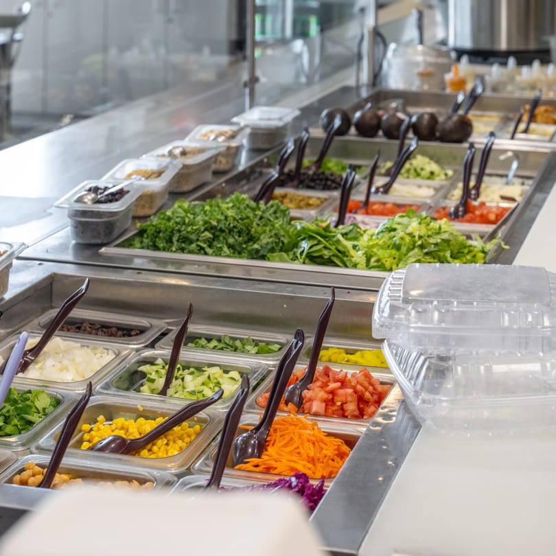 A salad bar at Natural Greens with various fresh ingredients in metal bins and serving tongs.
