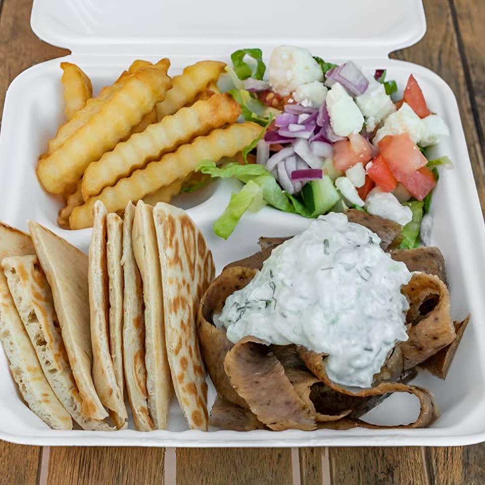 Gyro platter with meat, pita bread, fries, Greek salad, and tzatziki sauce in a takeout container.